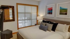 Een hotelkamer met een groot bed, een televisie, een raam met zonwering en twee kunstwerken in een lijst aan de muur.