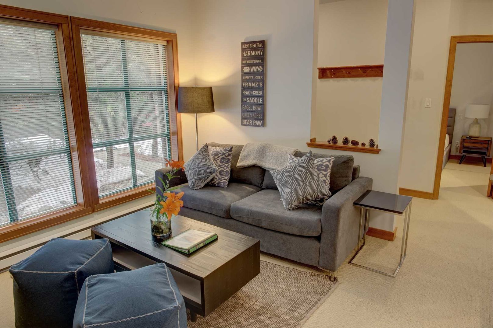 Ein Wohnzimmer mit grauem Sofa, zwei blauen Sesseln, einem Couchtisch und einer Lampe. Ein Fenster mit Jalousien, ein an der Wand befestigtes Schild und eine kleine Ablage mit Dekorationselementen sind ebenfalls vorhanden.