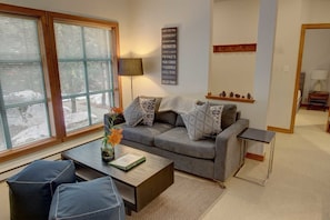 Una sala de estar con un sofá gris, dos sillones azules, una mesa de centro y una lámpara. Se aprecia una ventana con persianas, un letrero en la pared y un pequeño estante con objetos decorativos.