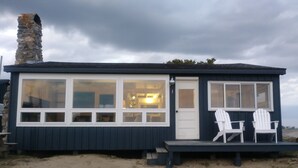 Una pequeña casa de playa de color oscuro con chimenea de piedra, ventanas grandes y dos sillas blancas en el porche.