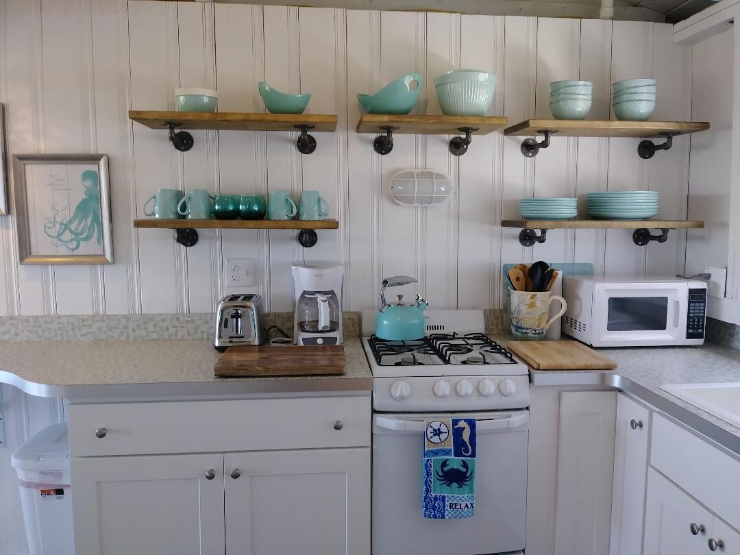 Une cuisine avec des meubles blancs, une cuisinière à gaz, un four à micro-ondes et de la vaisselle de couleur turquoise rangée sur des étagères.