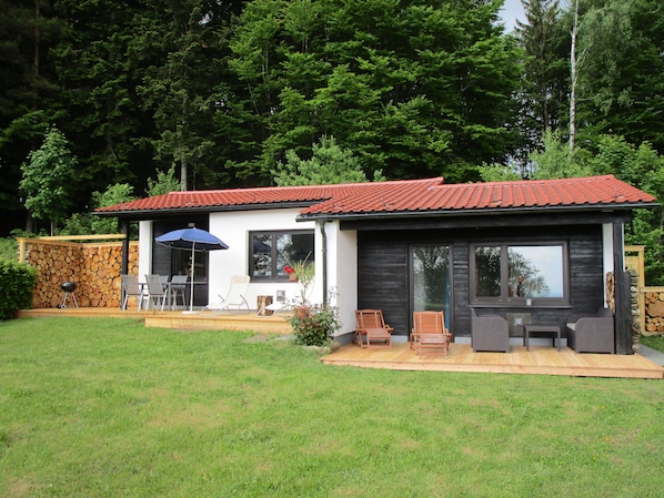 Une petite cabane à deux étages, avec un toit recouvert de tuiles rouges, une terrasse dotée de mobilier d’extérieur et un mur en pierre sur lequel sont empilés des troncs d’arbres.