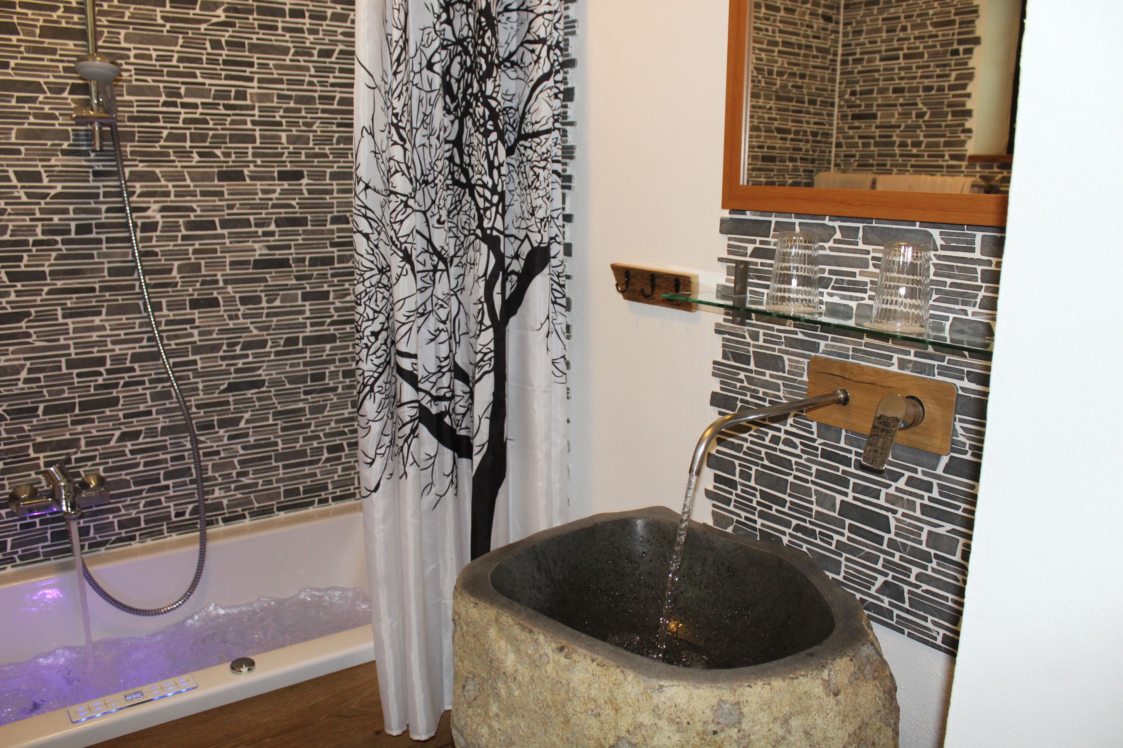 A bathroom with a stone sink, a shower with a tree-themed curtain, and a glass shelf with two glasses.