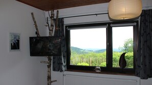 Ein Zimmer mit großem Fenster, Heizkörper, einem an einem Ast befestigten Fernseher und Blick ins Grüne.