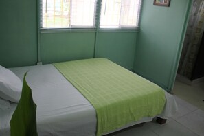 Un lit simple avec une couvre-lit verte, dans une pièce aux murs verts et dotée d’une fenêtre avec des stores.