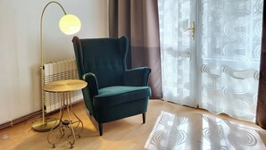 Ein Zimmer mit einem petrolfarbenen Sessel, einer Stehlampe, einem gemusterten Teppich und einem Fenster mit Vorhängen.