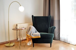 Ein Zimmer mit einem dunkelgrünen Sessel, einer Stehlampe, einem kleinen runden Tisch mit Büchern und Gläsern sowie einer Heizung.