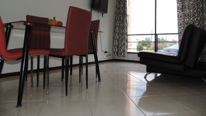 A dining area with a table and chairs, a television mounted on the wall, and a large window with patterned curtains. - Apartamento confortable (Bogotá)