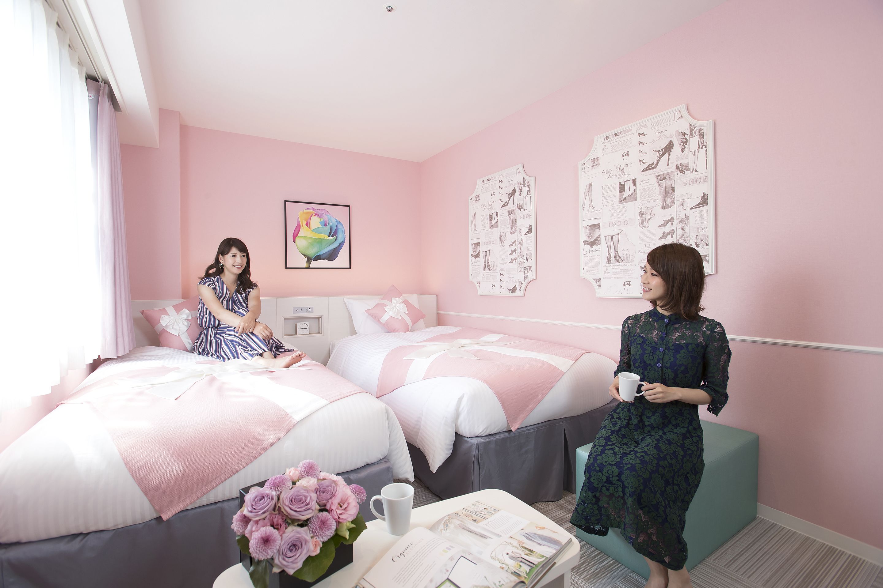 A pink-themed bedroom with two beds, a coffee table with a vase of flowers, and a wall with fashion posters.