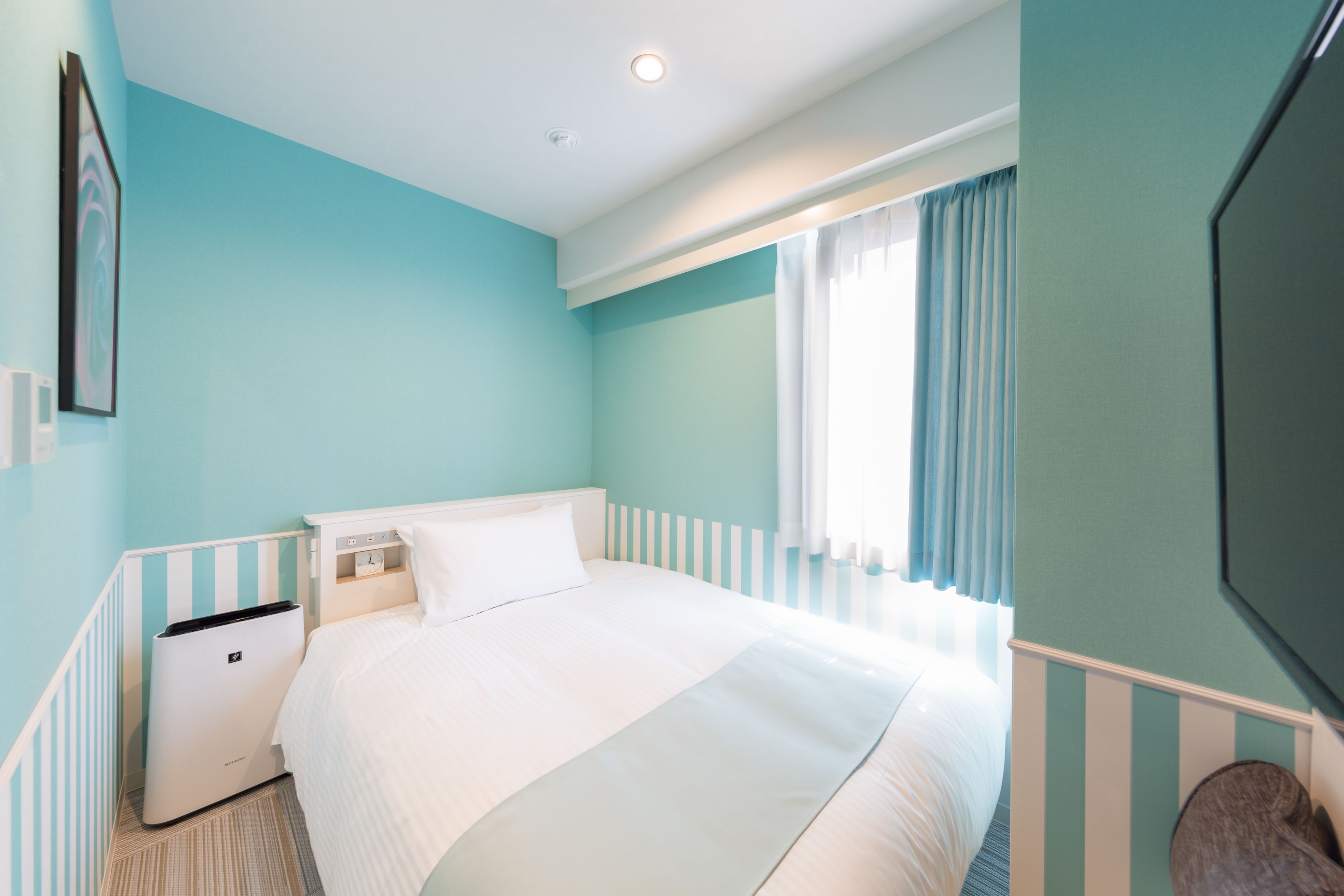 A hotel room with a bed, a bedside table, a trash bin, a window with curtains, and a wall-mounted TV.