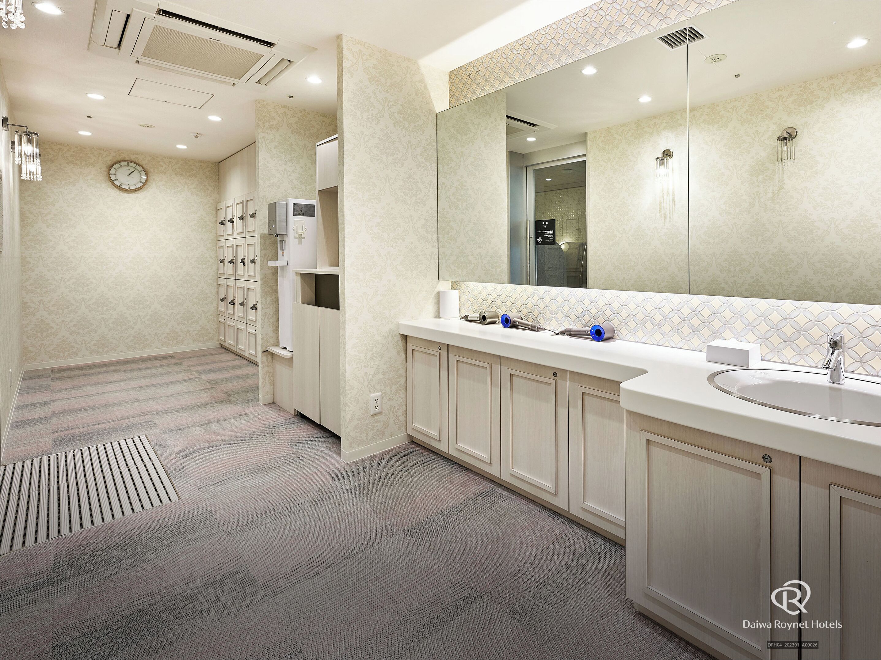 A modern bathroom with a large mirror, a sink, and a shower. There are towel dispensers and a clock on the wall.