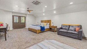 A bedroom with a bed, a wooden headboard, a gray leather couch, a wooden side table, and a ceiling fan. - Cozy Studio • Trail Access • Walk to Payette Lake (McCall)