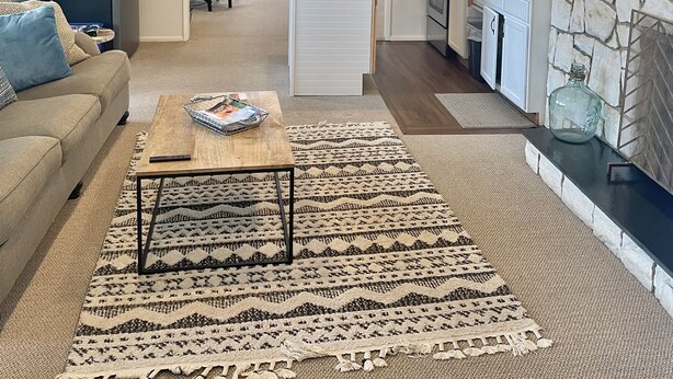 A modern living room with a sofa, coffee table, and patterned rug. A kitchen with a stone backsplash and a dining area with a table and chairs are visible in the background.