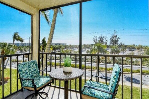 A balcony with two chairs and a small table, overlooking a street with palm trees and buildings.