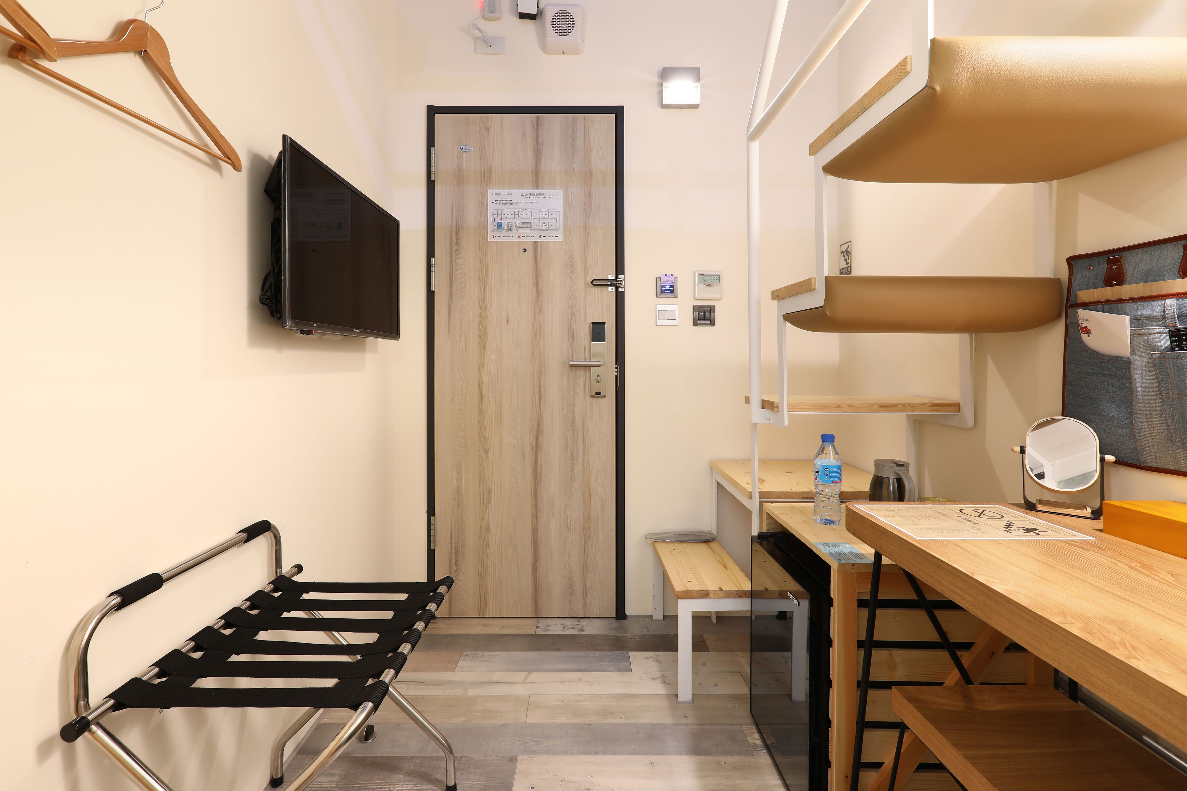 A hotel room with a wooden door, a flat-screen TV mounted on the wall, a wooden bench, a small table with a water bottle, and a mirror on the table.