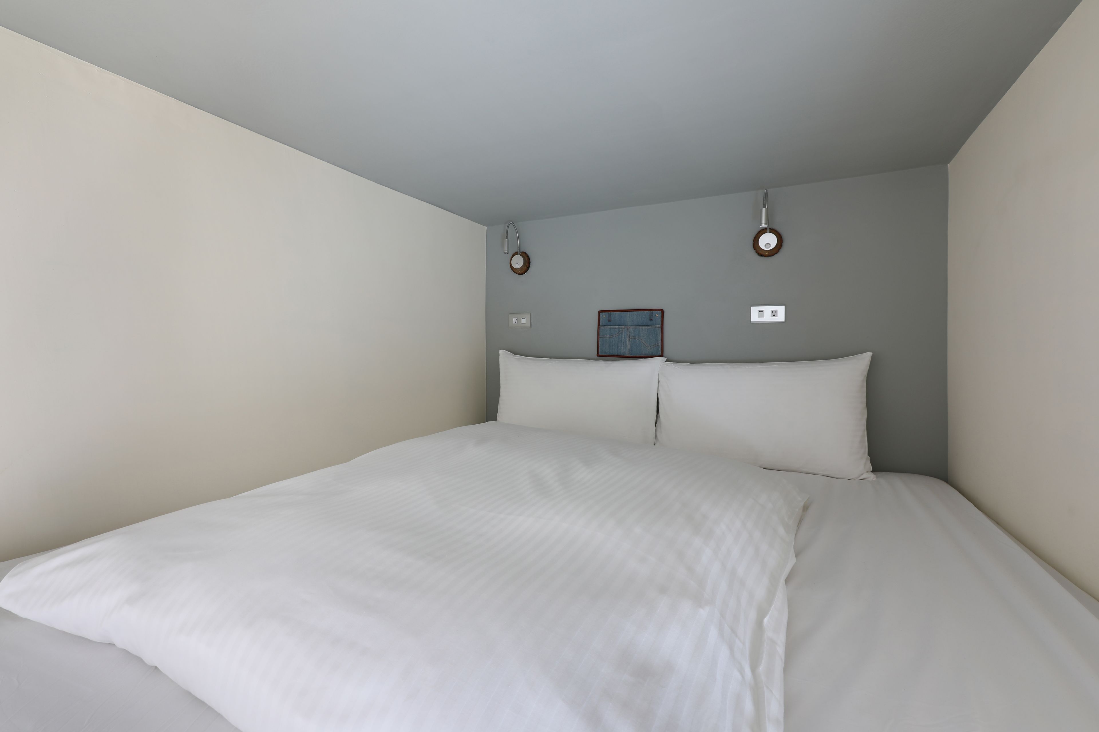 Cama bem arrumada em quarto com paredes cinza claro, duas luminárias fixadas na parede e uma pequena moldura com imagem na parede.