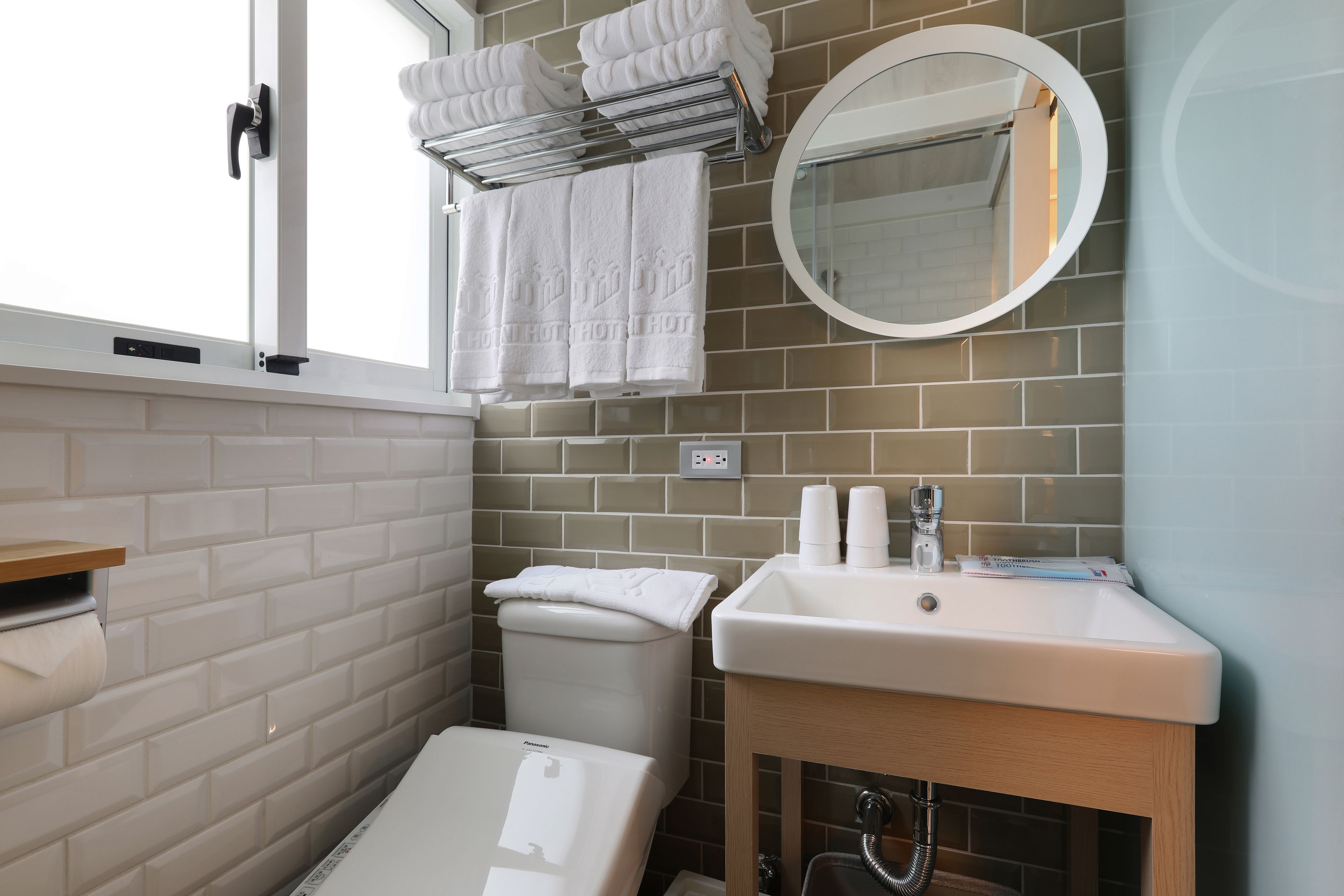 Un baño con paredes de azulejos blancos, un lavabo blanco, un espejo y un perchero con toallas dobladas.