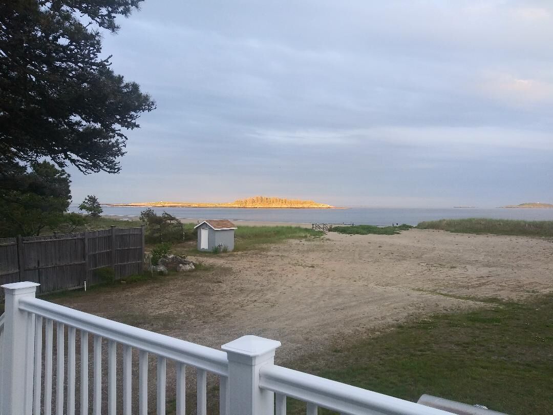 Una spiaggia sabbiosa con una recinzione di legno e un piccolo deposito, affacciata su una distesa d'acqua e una terraferma in lontananza.