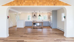 Cocina amplia con piso de madera, una isla central, iluminación colgante y vistas al exterior a través de ventanas grandes.