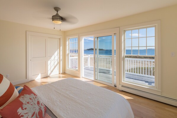 A bedroom with a large bed, a ceiling fan, and a view of a beach through the sliding glass doors.