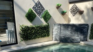 Una piscina con un muro de piedra y paneles decorativos, rodeada de follaje y un cerramiento de vidrio transparente.