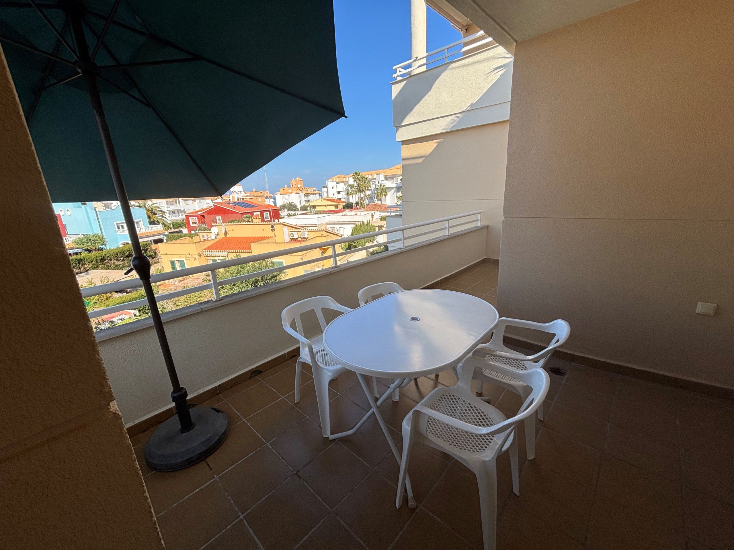 Een balkon met een witte tafel en stoelen, een groene parasol en uitzicht op gebouwen en groen.