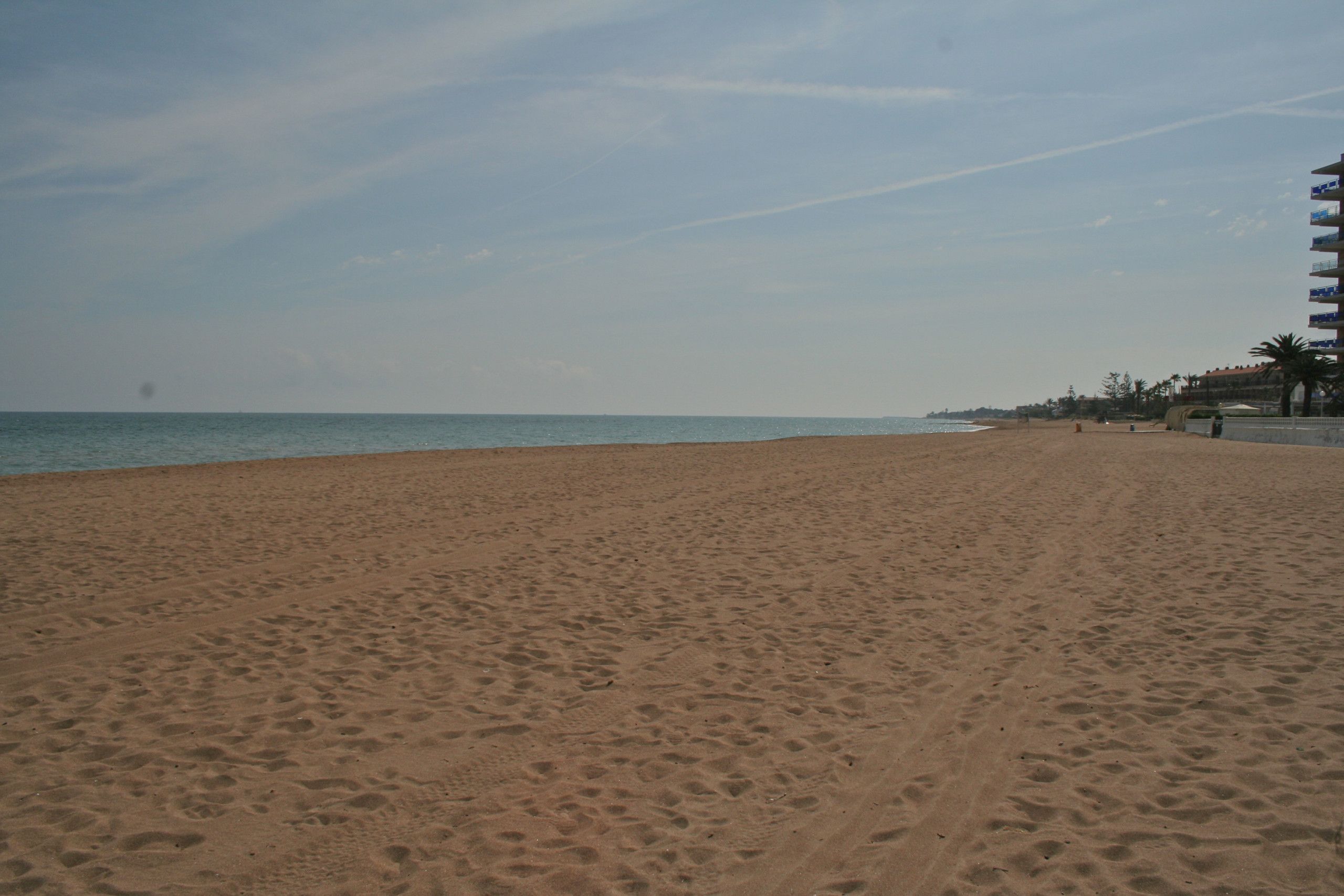 Une vaste plage de sable où l’on peut voir des empreintes de pas menant à l’eau, un ciel dégagé et, à droite, un bâtiment doté de nombreux balcons.