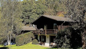 A log cabin with a porch, surrounded by dense trees and a grassy area. - Haus 2  - Typ C (kombiniert) (Schönecken)