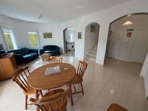 En rummelig opholdsstue med et rundt træbord, flere træstole, en sofa og en sofalounge. Der er en trappe til en etage ovenover, en dør med teksten 'SALIDA', og en vaskemaskine i baggrunden.