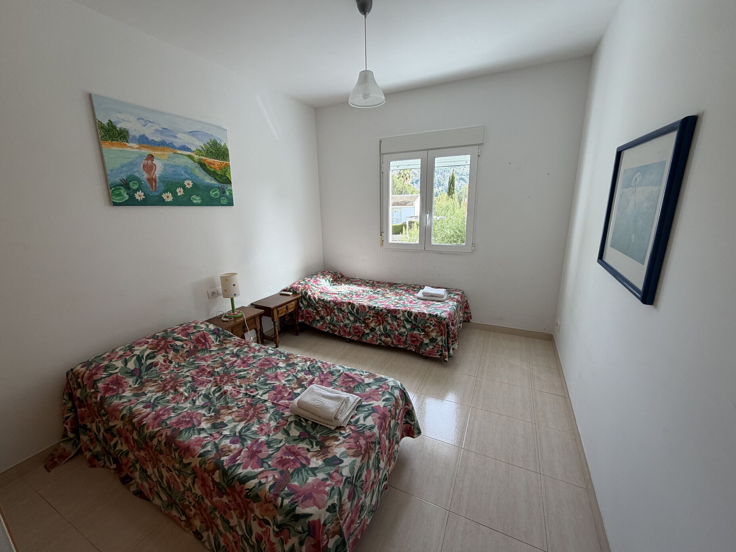 A bedroom with two beds, a desk, a lamp, a window, and a painting on the wall.