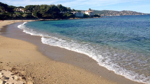A beach with clear blue water, sandy shore, and houses in the background.