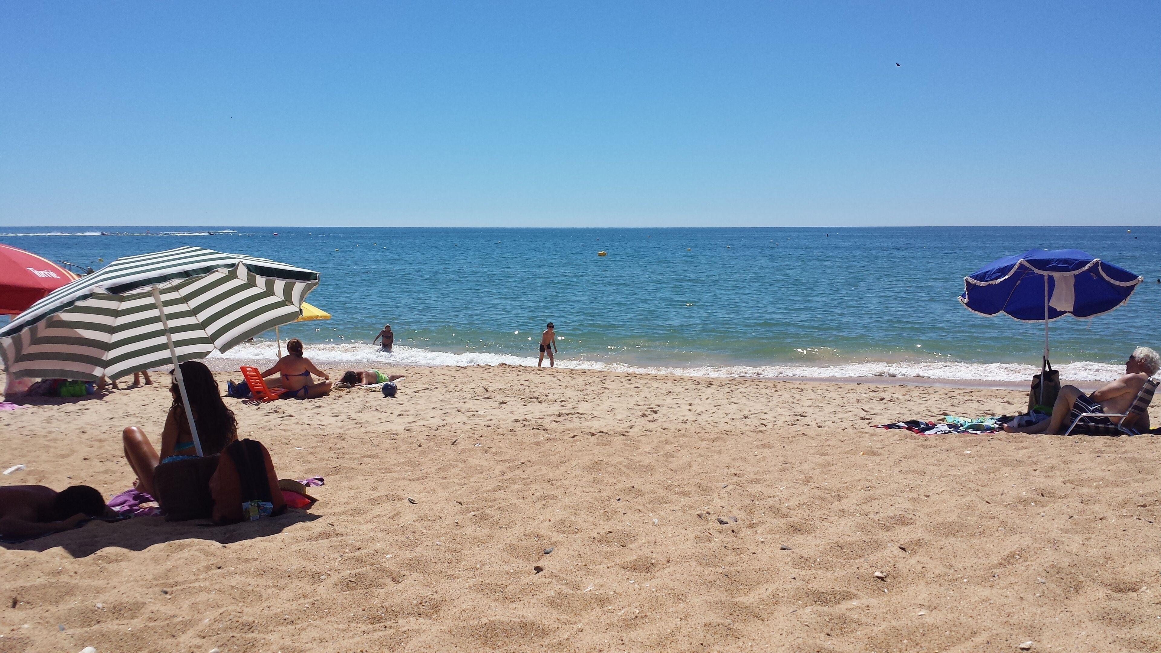 Una platja amb gent relaxant-se sota ombrelles, un cel serè i l'oceà al fons.