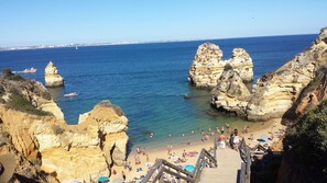 A coastal beach with rocky cliffs, clear blue water, and a wooden staircase leading to the beach.