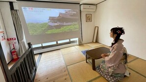 A room with tatami flooring, a projection screen displaying a building, and a fire extinguisher on the wall. - Guest house Connection Karasuma Gojo (Kyoto)