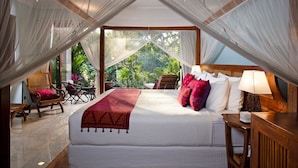 A bedroom with a large bed, a bedside table with a lamp, and a view of lush greenery outside. - Kano Sari (Ubud)