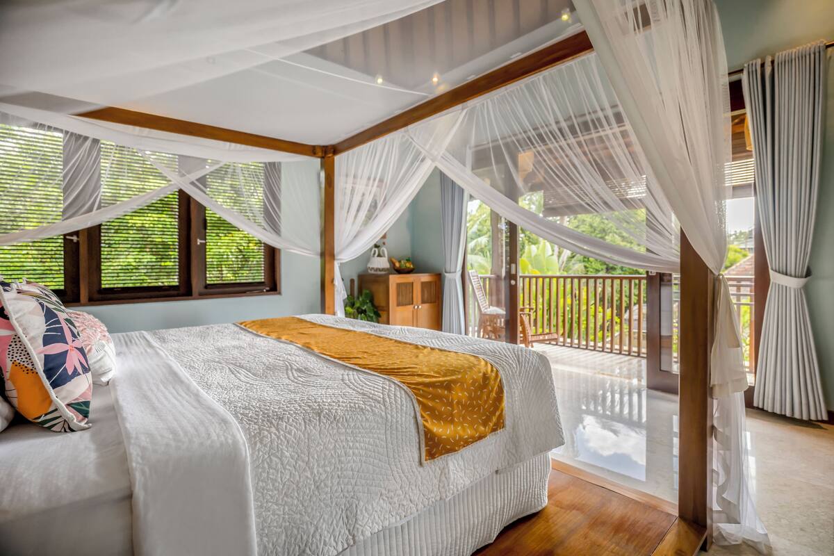 A bedroom with a large bed, a wooden floor, a balcony with a view of greenery, and a window with sheer curtains.