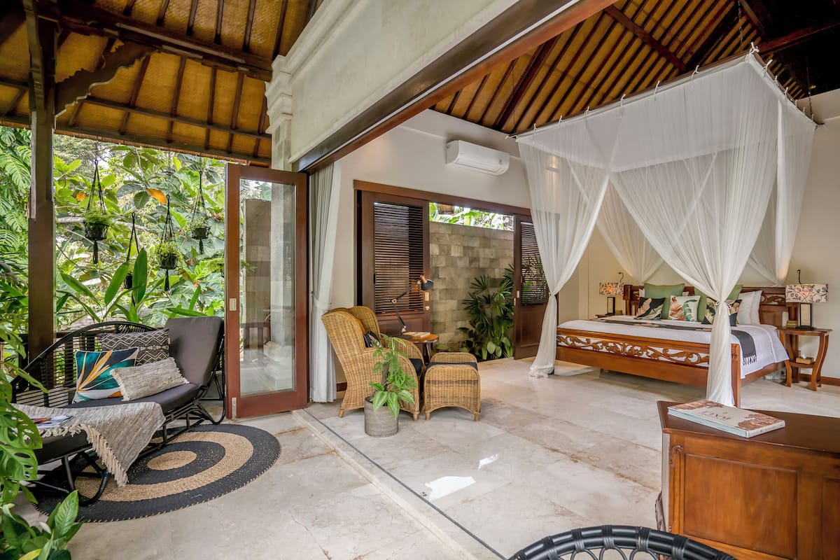 A room with a bed, a sitting area with a chair and a table, and a large window with a view of greenery.