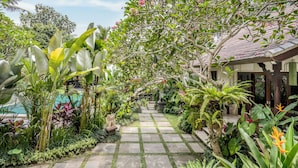 A garden pathway leading to a building with a tiled roof, surrounded by lush tropical plants and a swimming pool. - Kano Sari (Ubud)