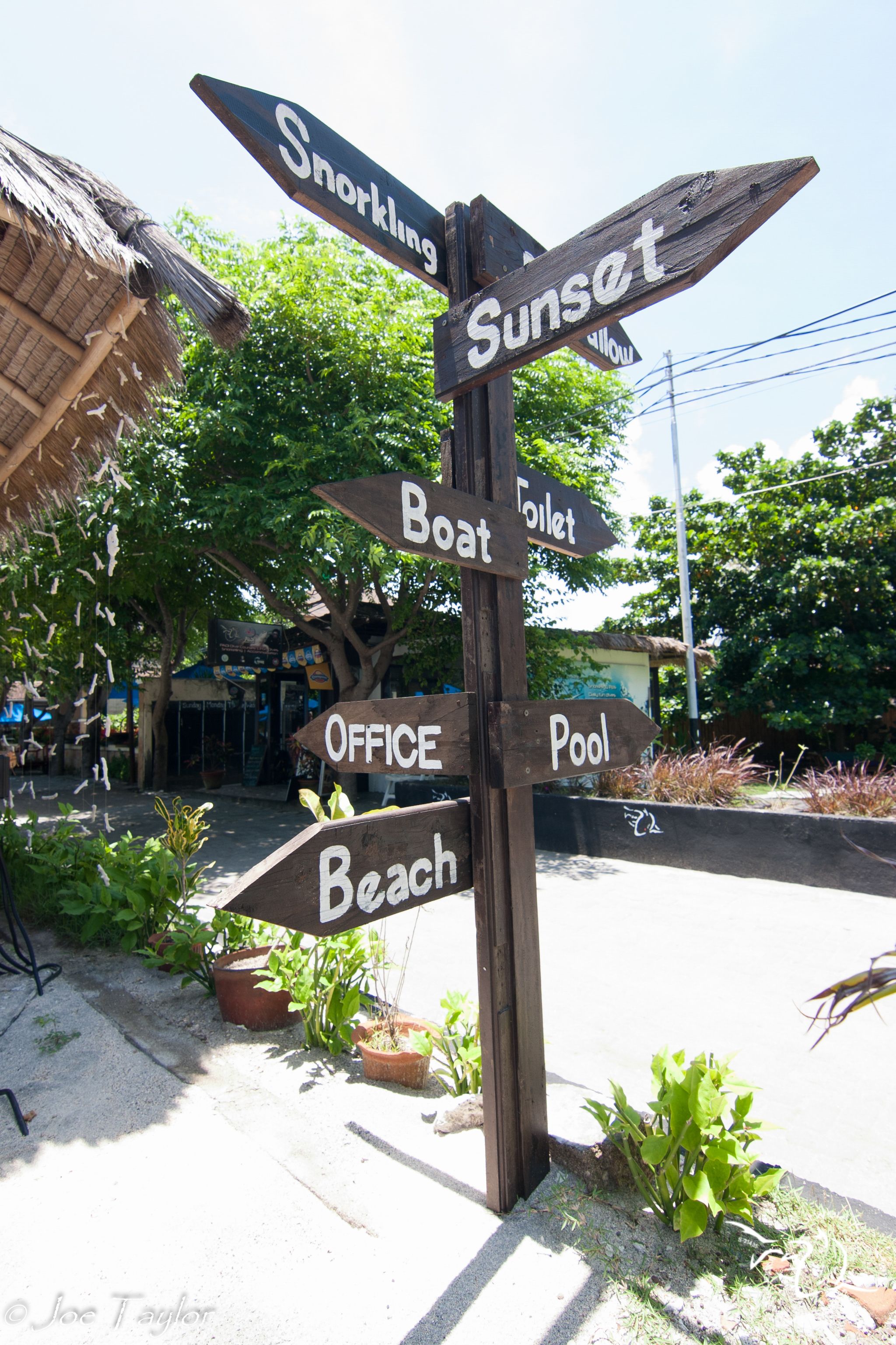 A wooden signpost with multiple directional signs pointing to various amenities such as 'Snorkeling', 'Sunset', 'Boat', 'Toilet', 'Office', 'Pool', and 'Beach'.