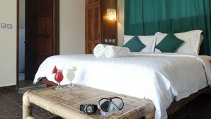 A neatly made bed with white linens, a wooden wardrobe, a table with a drink and sunglasses, and a green curtain.