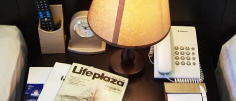 A bedside table with a lamp, a remote control holder, a clock, a telephone, and a Lifeplaza magazine.