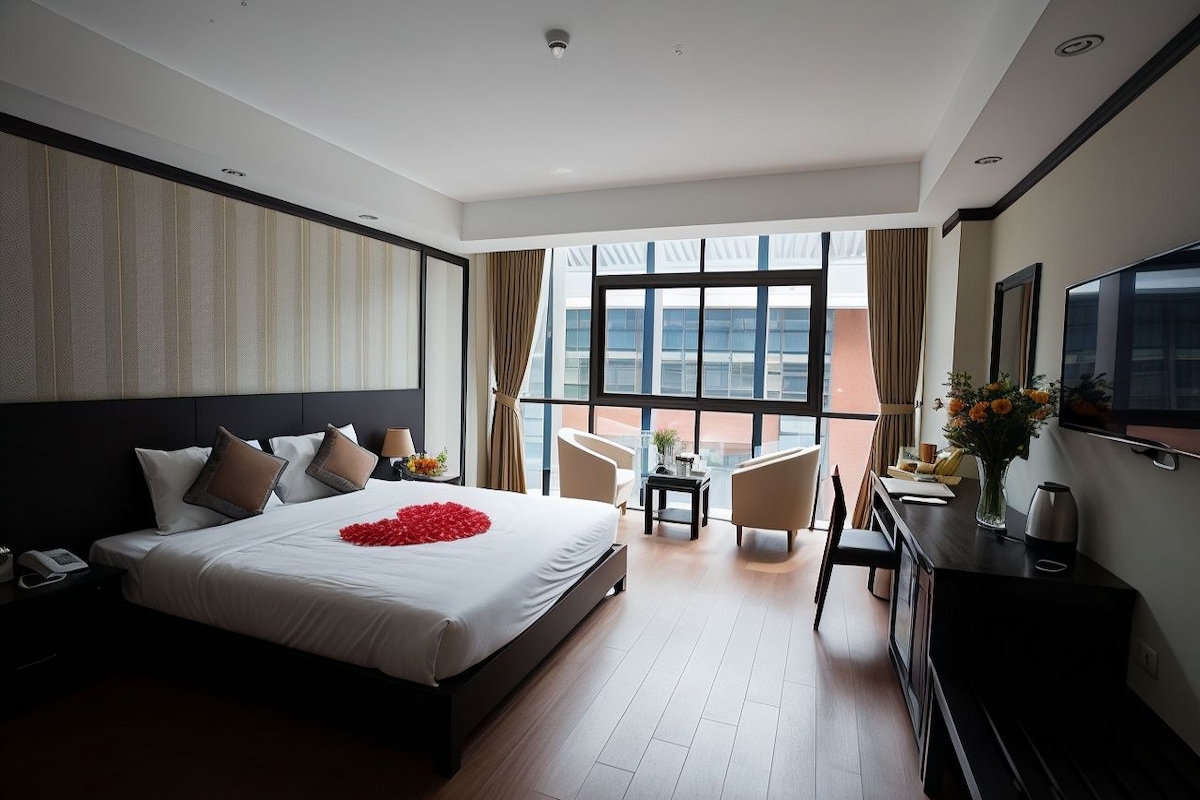 A hotel room with a large bed, a desk, a chair, a TV, and a view of the city through large windows.
