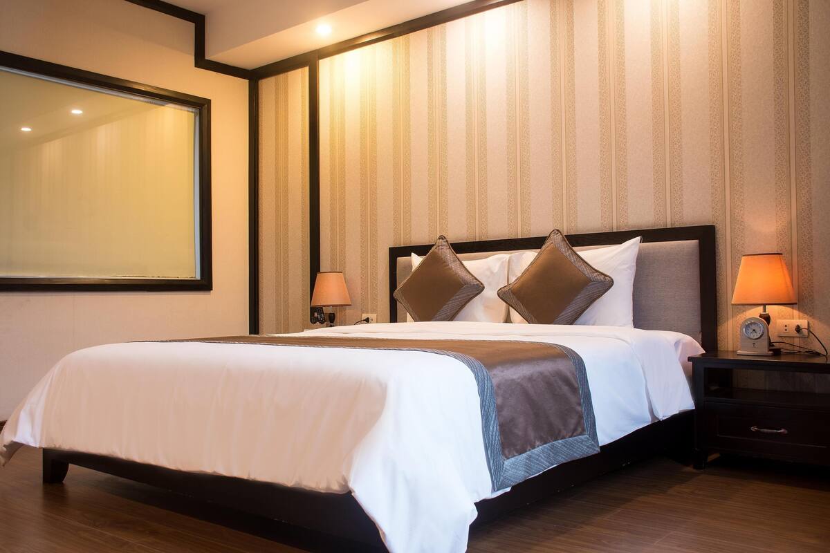 Hotellihuone, jossa on suuri sänky, kaksi yöpöytää, yölamppu ja seinään kiinnitetty valaisin.
