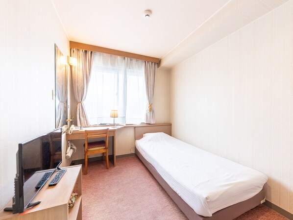 Quarto de hotel com uma cama de solteiro, uma escrivaninha com cadeira, televisão e janela com cortinas.
