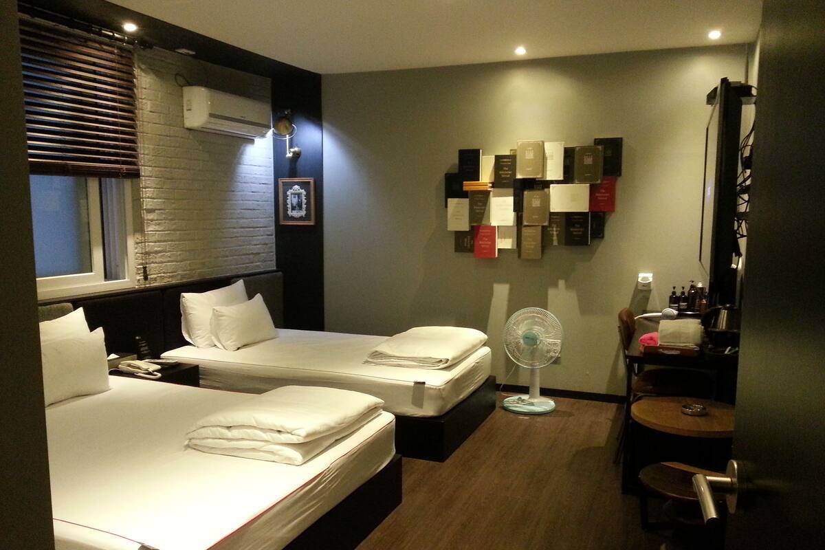 A hotel room with two beds, a desk, a chair, a fan, and a wall with framed pictures and books.