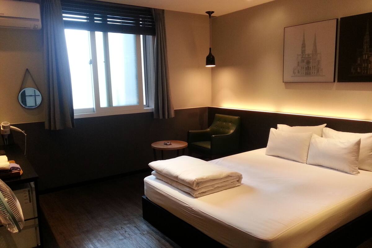A hotel room with a large bed, a chair, a small table, and a window with curtains.