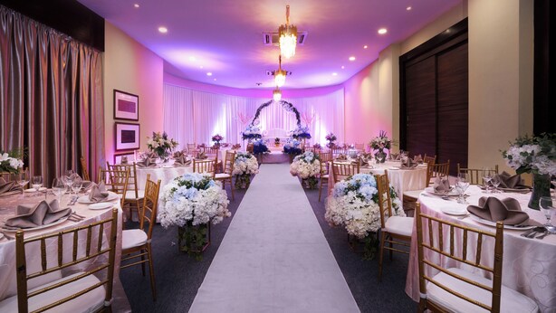 Un salón de banquetes con un pasillo central, mesas dispuestas para cenar y un arco decorativo al final.