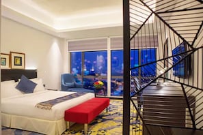 Habitación de hotel con cama, un banco rojo, una silla azul y vista de la ciudad a través de amplias ventanas.