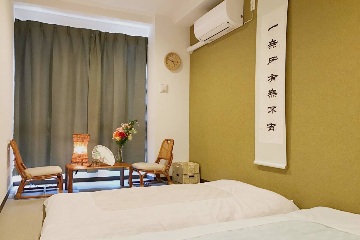 A hotel room with a bed, two chairs, a table with a lamp, a clock, and a wall-mounted air conditioner.