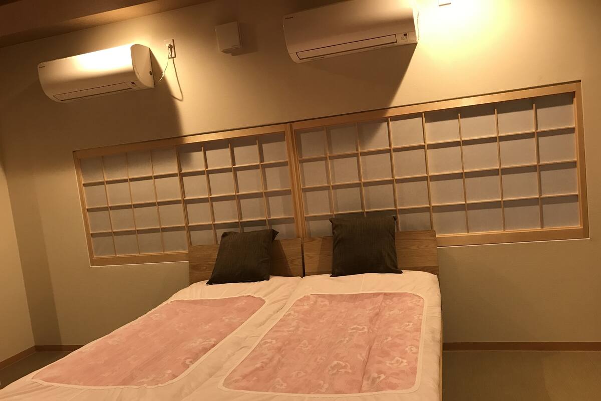 A bedroom with a bed, two pillows, a window with a wooden frame, and two wall-mounted air conditioners.